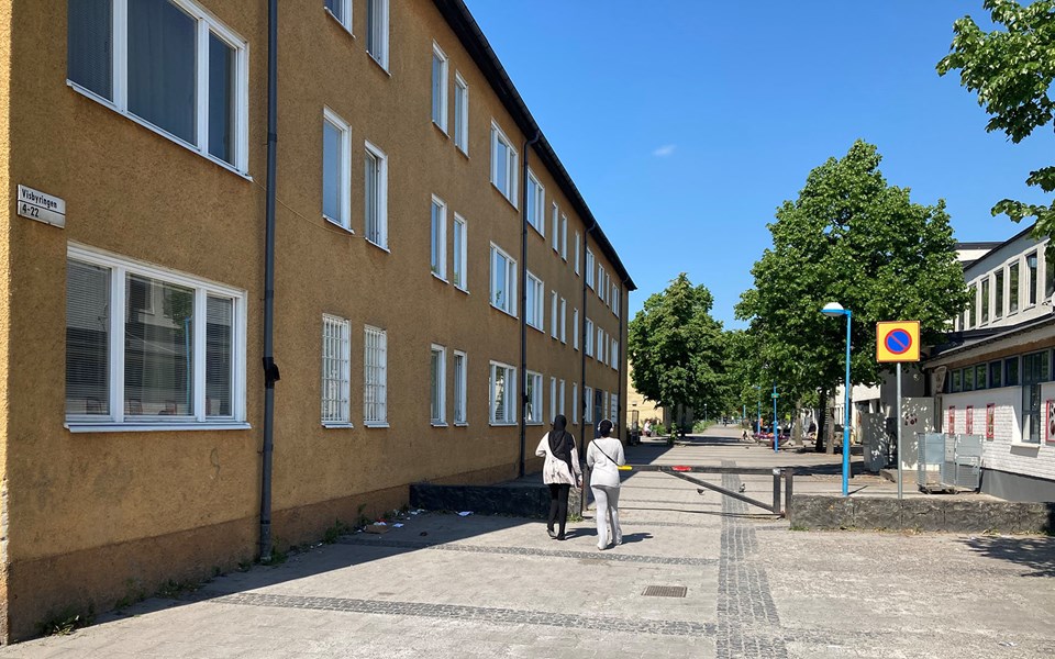 Gångstråket Rinkebygången omgiven av byggnader. Två personer promenerar. Längre fram längs gatan syns träd